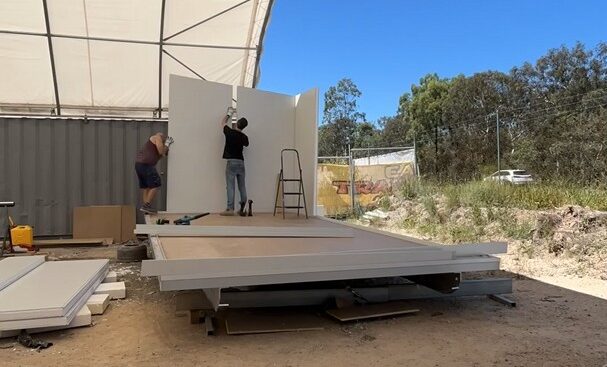 Dan and Ancel stand on the base of a semi-constructed tiny home, putting up wall panels